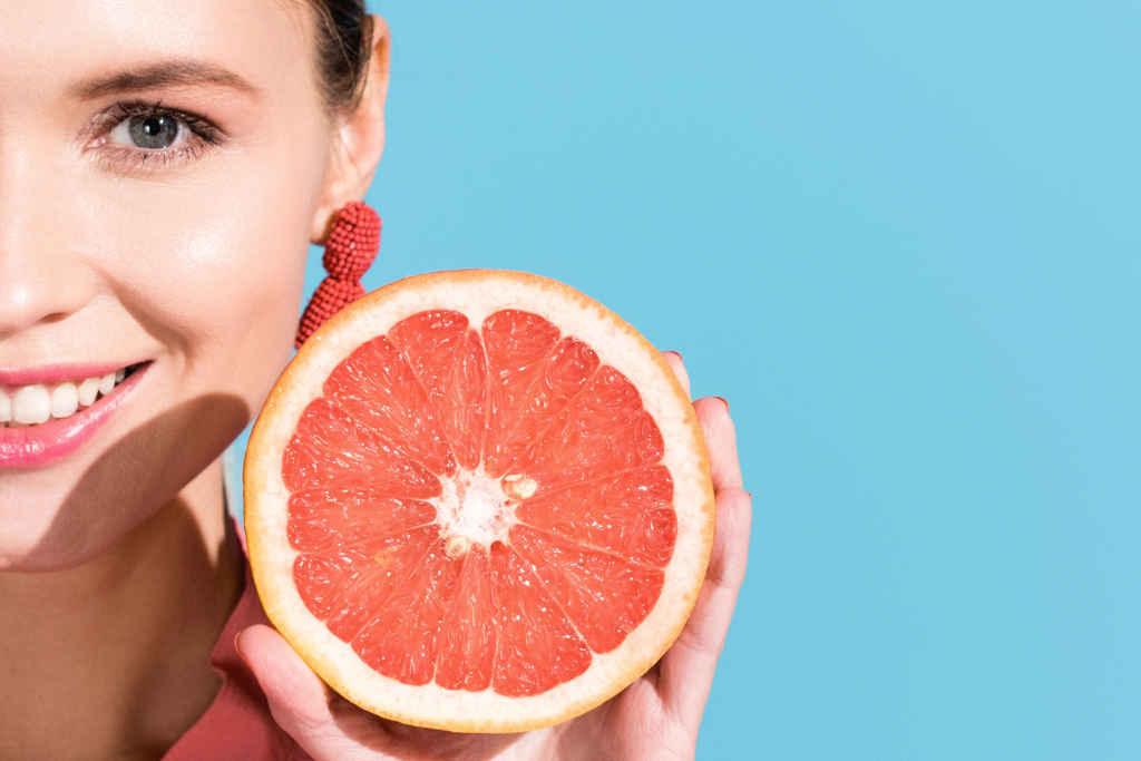 Uśmiechnięta kobieta trzymająca połówkę grejpfruta na tle jasnoniebieskiego tła; zbliżenie na zdrowe zęby i owoc o intensywnym, różowym miąższu